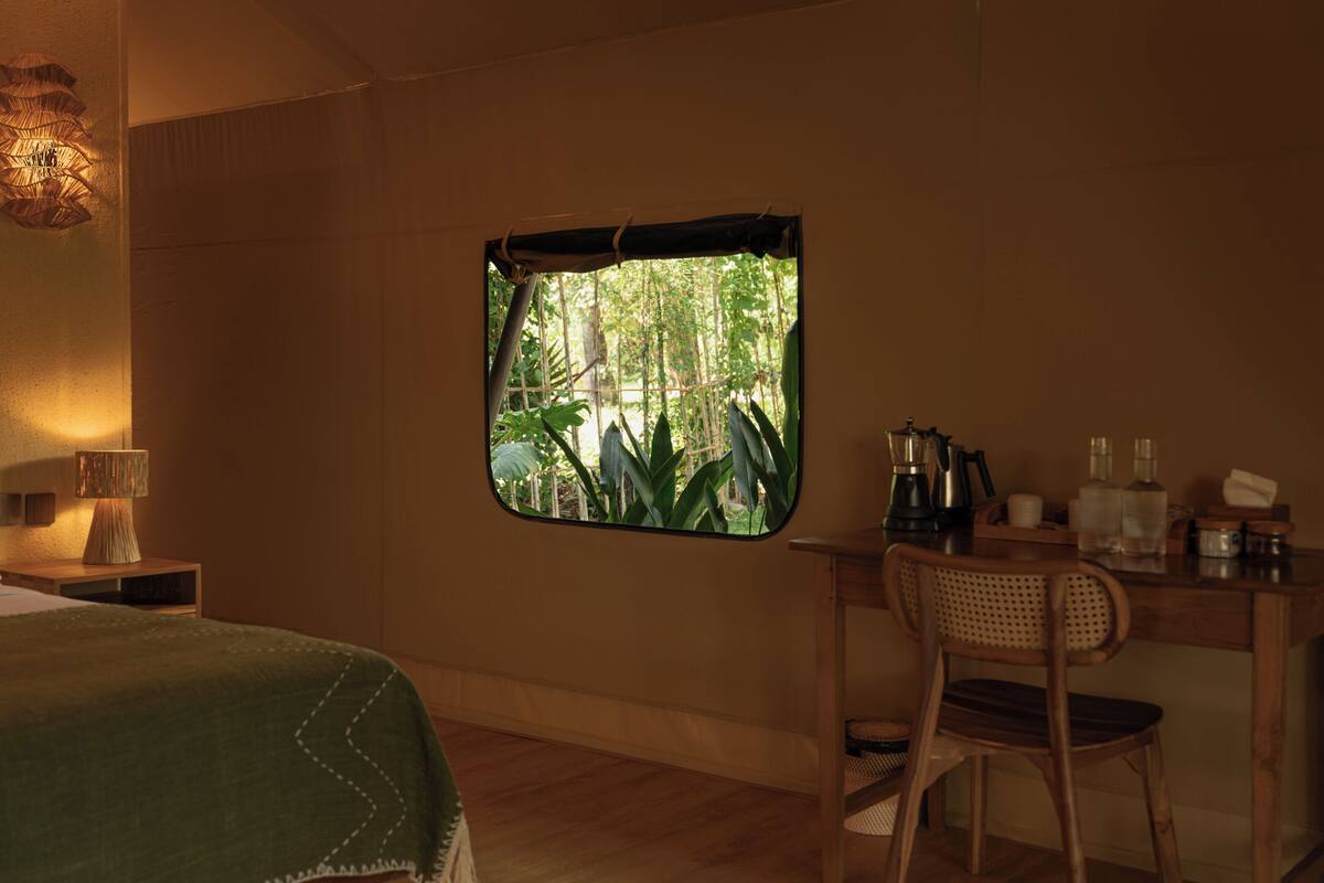 Ein Zimmer mit Bett, Schreibtisch mit Stuhl, Fenster mit Blick auf Grünflächen und einer Lampe auf dem Nachttisch.