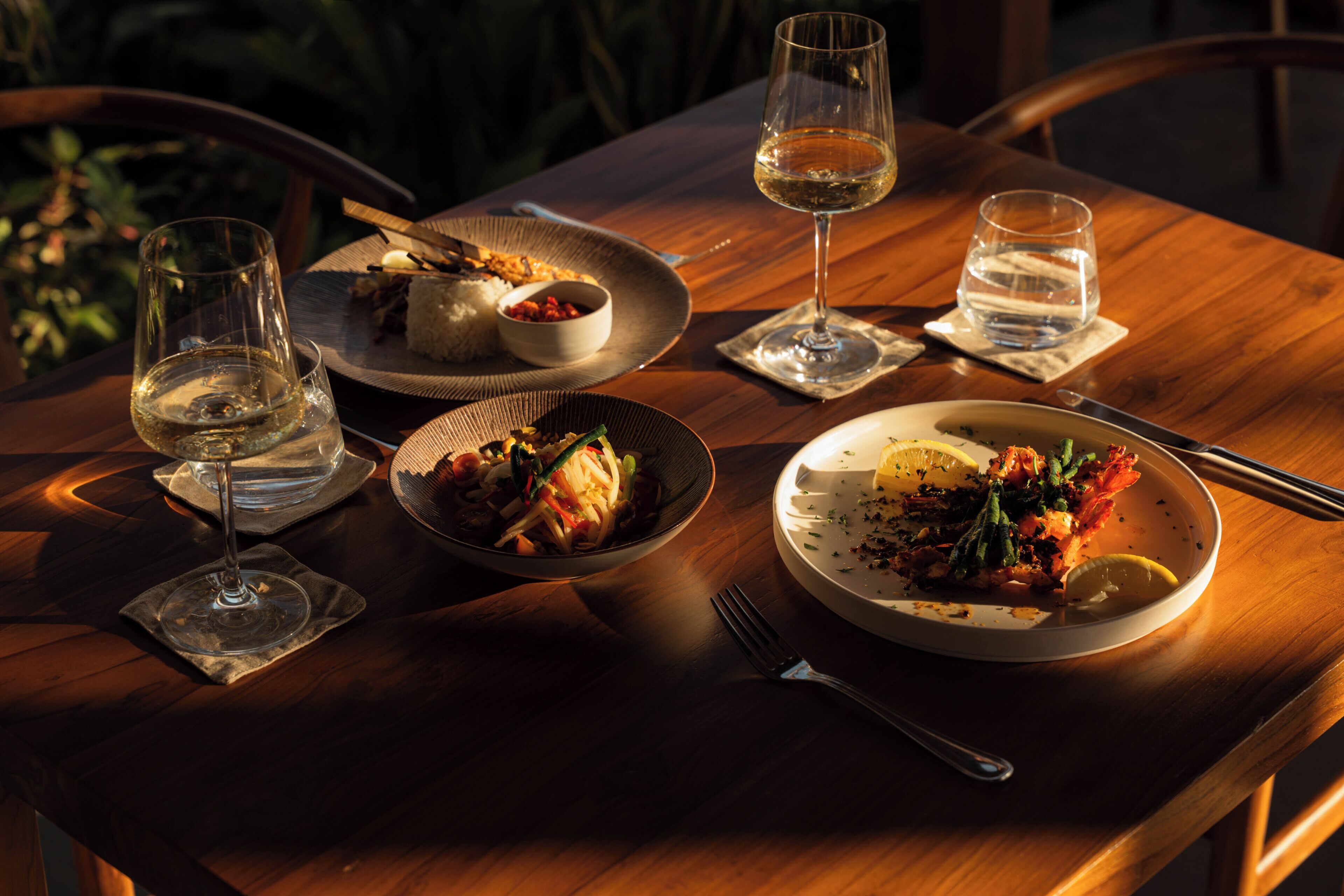 A wooden dining table with plates of food, glasses of white wine, and cutlery.