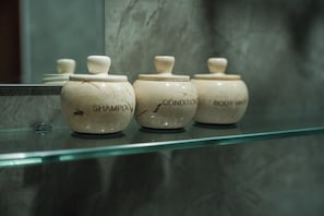 Three ceramic jars labeled 'SHAMPOO,' 'CONDITIONER,' and 'BODY WASH' on a glass shelf. - Vela Tianyar Boutique Resort By Dhananjaya Hospitality (Kubu)