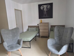 A room with two grey armchairs, a bed with a green bedspread, a wooden dresser, and a wall-mounted elephant artwork.