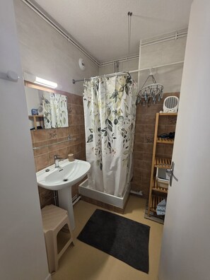 A bathroom with a white sink, a shower curtain with a leaf pattern, a small stool, and a black rug.