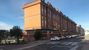 Un largo edificio de ladrillo rojo con numerosas ventanas y techo plano, situado junto a una carretera con coches estacionados.