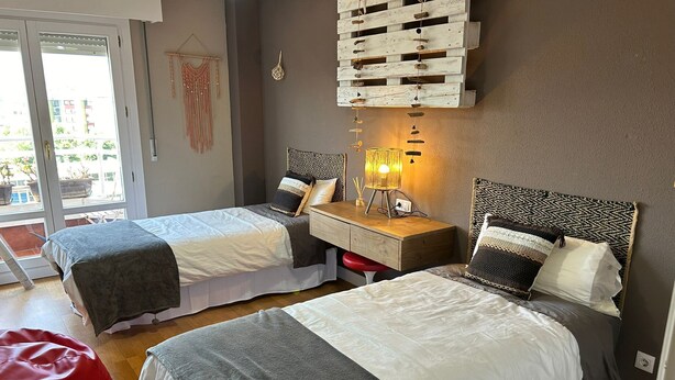 Un dormitorio con dos camas, una mesita de noche de madera, un sillón de peluche rojo y una lámpara fijada a la pared.