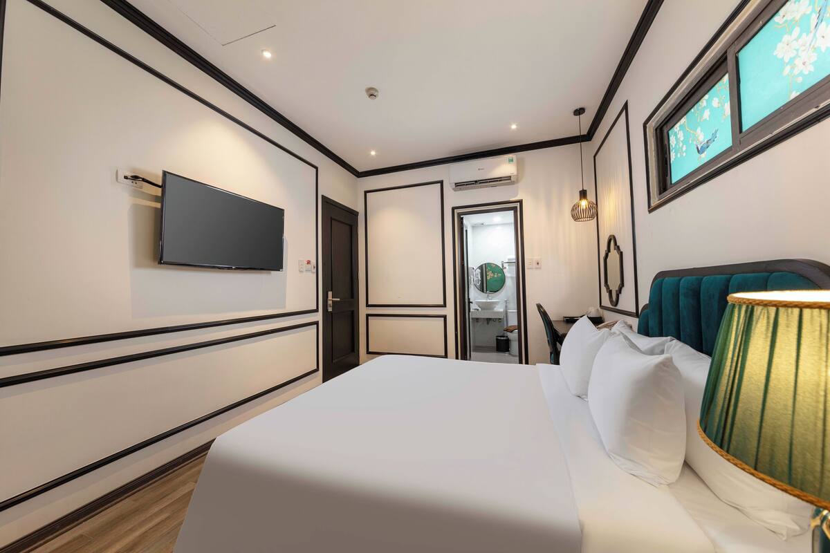 A hotel room with a bed, a flat-screen TV mounted on the wall, a window with a view, and a door leading to a bathroom.
