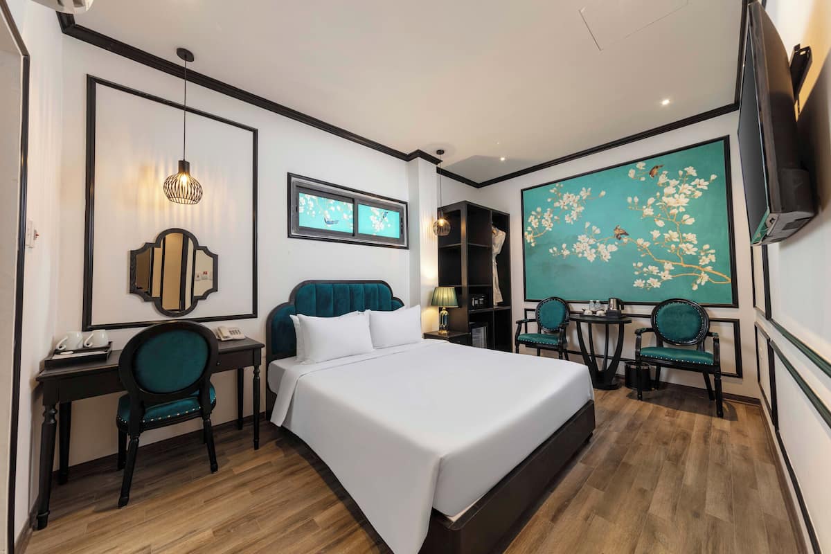 A hotel room with a large bed, two chairs, a desk, a TV, and a decorative wall painting.