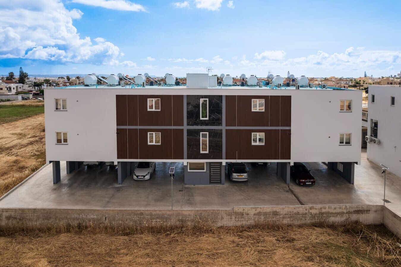 A modern multi-story building with a flat roof, parking spaces, and a flat landscape in the background.