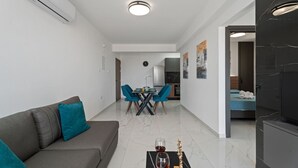 Un salon moderne avec un canapé gris, un coin repas comprenant une table et des chaises, un téléviseur et une cuisine visible à travers une porte ouverte.