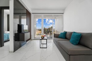 Ein modernes Wohnzimmer mit einer Couch, einem Couchtisch und einem Fernseher. Es gibt einen Balkon mit Blick auf die Stadt.