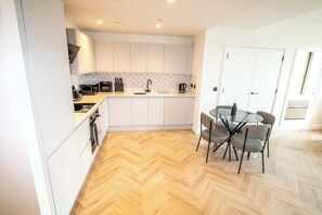 A modern kitchen with white cabinets, a herringbone wood floor, and a dining area with a glass table and chairs.