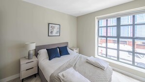 Une chambre moderne avec une grande fenêtre, un lit recouvert de linge de lit blanc, une table de chevet et un tableau accroché au mur.
