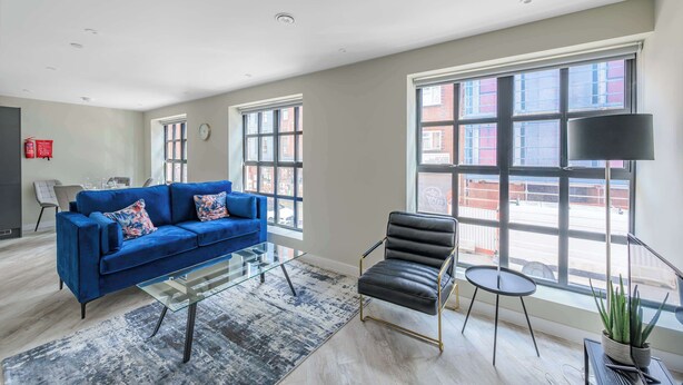 Un salon moderne avec un canapé bleu, un fauteuil en cuir noir, une table basse en verre et de grandes fenêtres donnant sur la rue.