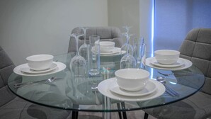Une table ronde en verre, dressée avec de l’assiette blanche, des bols et des couverts, entourée de chaises grises à revêtement en tissu, dans une pièce moderne aux couleurs neutres.