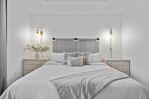 A bedroom with a bed, two bedside tables, a nightstand, a headboard, and two wall-mounted lamps.