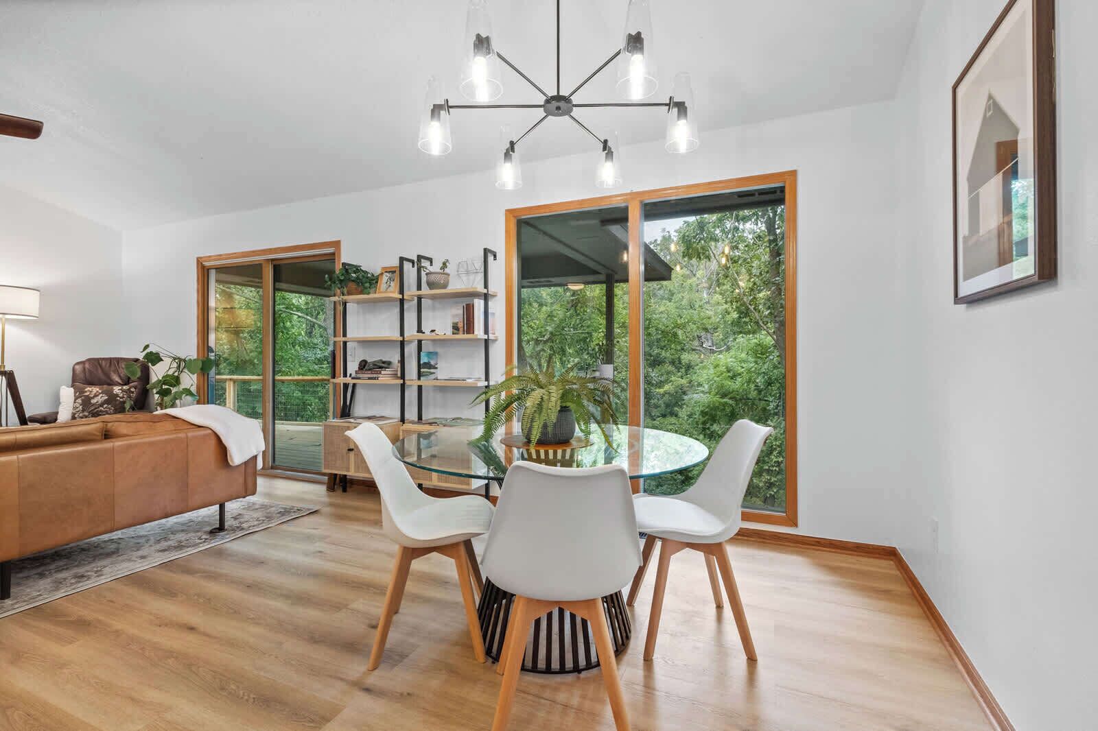 A modern living room with a glass dining table, white chairs, a leather sofa, a bookshelf, and a large window with a view of greenery.