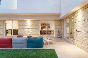 A modern outdoor living space with a dining area, a kitchen visible through glass doors, and a stone-textured wall. - Villa Bel Prato with pool - My Rental Homes (Modica)