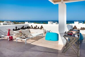 A rooftop terrace with white walls, hexagonal patterns, and a view of the sea. There are lounge chairs, a table with drinks, and a blue chair. - Villa Matilde with sea view - My Rental Homes (Noto)