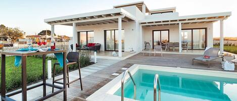 A modern outdoor pool area with a wooden deck, lounge chairs, and a dining setup.