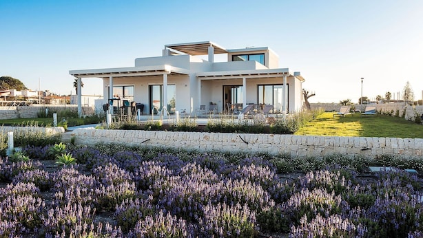 A modern two-story house with a patio area and a garden with purple flowers. - Villa Orietta, garden and pool - My Rental Homes (Noto)