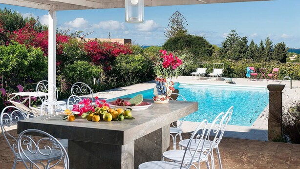 Zona junto a la piscina con una mesa dispuesta para comer, rodeada de sillas de metal blanco.