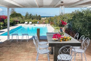 A poolside area with a view of the sea, a table with fruits and flowers, and white metal chairs. - The Flower Garden with pool - My Rental Homes (Marsala)