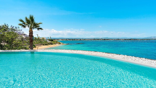 A clear blue pool with a white pebble border, a palm tree, and a view of the sea and distant mountains. - Sea Art Villa with private pool - My Rental Homes (Syracuse)