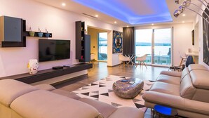 A modern living room with a flat-screen TV, comfortable seating, a glass table, and a view of the sea. - Sea Art Villa with private pool - My Rental Homes (Syracuse)
