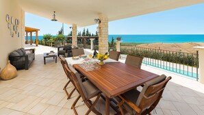 Eine Terrasse mit Holztisch und -stühlen, einer Couch und einer Ziervase. Im Hintergrund ist Meerblick und ein Pool zu sehen.