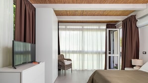 Een moderne slaapkamer met een flatscreen televisie, een rieten stoel, een bed met een beige deken en een groot raam met dunne gordijnen.