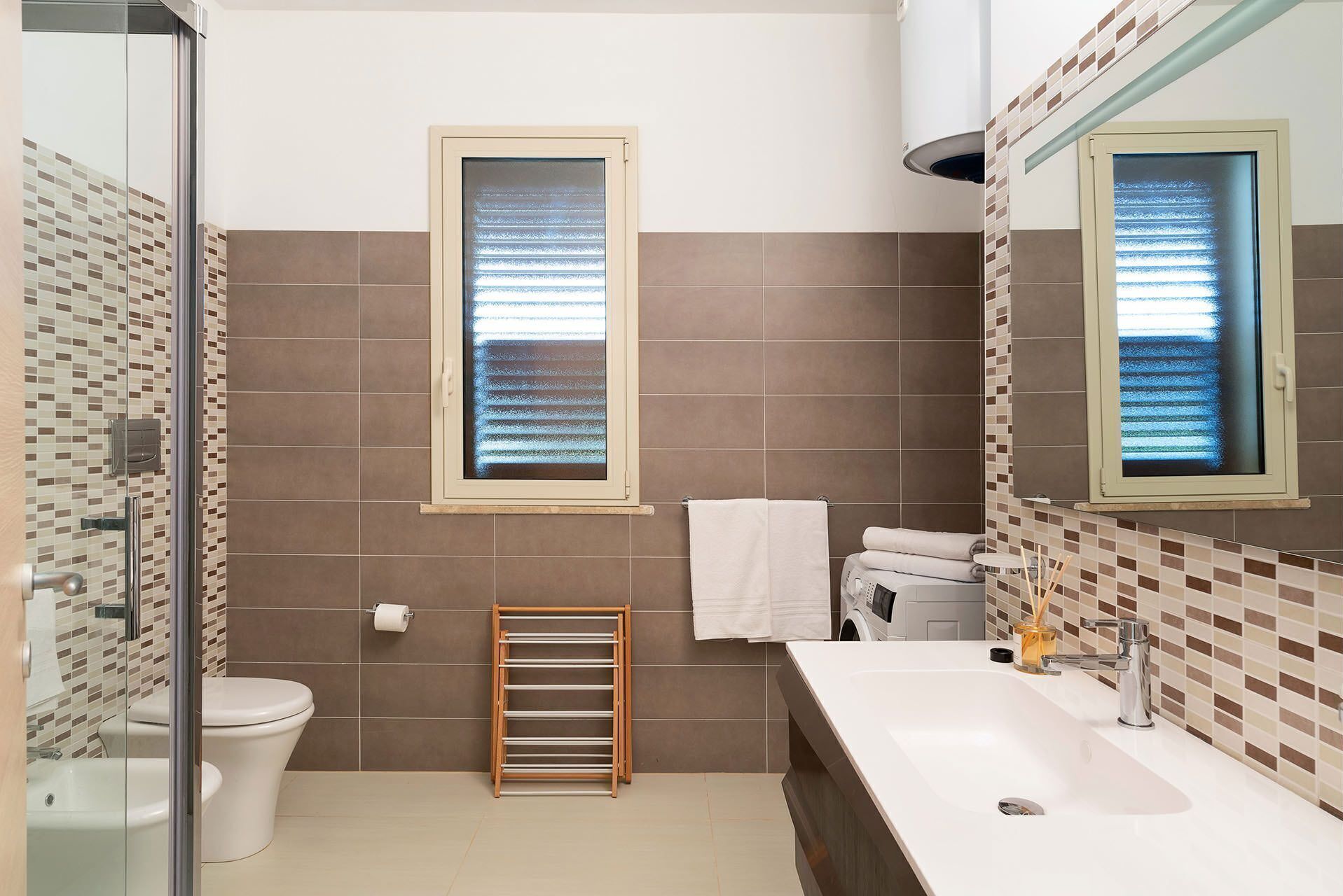 A bathroom with a toilet, bidet, and a sink with a mirror. There are two windows with blinds, a towel rack, and a towel hanging on the wall.