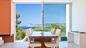 A modern dining area with a large table and chairs, a hanging light fixture, and a view of the sea and hills. - My Rental Homes - Villa Twilight with private pool and sea view (San Jose)
