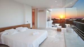 A modern hotel room with a large bed, a balcony with a city view, and a sunset visible through the window. - My Rental Homes - Villa Twilight with private pool and sea view (San Jose)