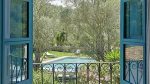 An open balcony with a view of a swimming pool, surrounded by lush greenery and a stone wall.