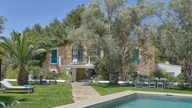 A poolside view of a stone house with a pool, surrounded by lush greenery and palm trees.
