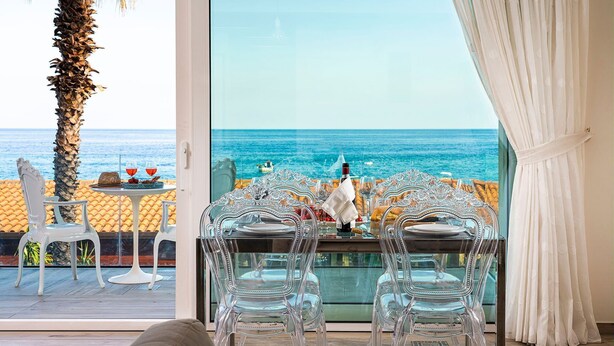 A dining area with a table set for two, clear chairs, and a view of the sea through a large window. - Casa Light Blu sea front home - My Rental Homes (Letojanni)
