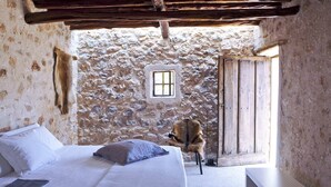 Une chambre rustique avec un mur en pierre, un lit recouvert de draps blancs, une petite fenêtre et une porte en bois.