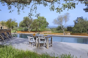 Área à beira da piscina com mesa e cadeiras, cercada por vegetação e um muro de pedra.