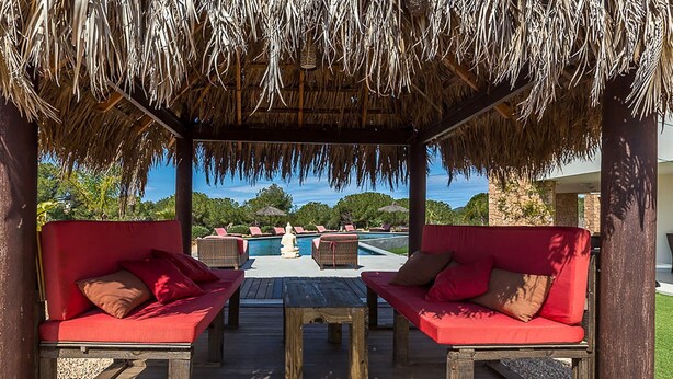A covered outdoor seating area with red cushioned sofas and a wooden table, overlooking a pool and lush greenery.