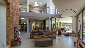 A spacious living area with a sectional sofa, a coffee table, and a fireplace. There is a staircase leading to an upper level, and large windows providing natural light.