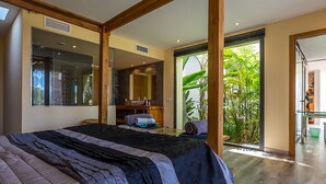A modern bedroom with a large bed, a bathroom with a mirror, and a sliding glass door leading to a balcony with plants.