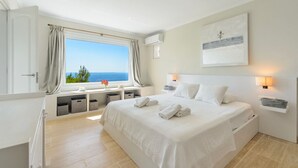 Un dormitorio con una cama grande, una ventana con vista al mar, una mesita de noche con lámpara y aire acondicionado instalado en la pared.