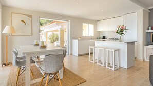 Una cocina moderna con armarios blancos, un comedor con mesa y sillas blancas, y una pequeña isla con taburetes.