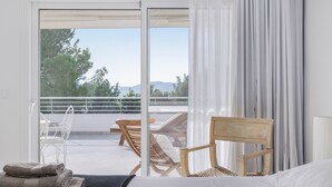 Hotelkamer met een bed, een houten stoel, een tafel met stoelen, en een balkon met uitzicht op bomen en bergen.