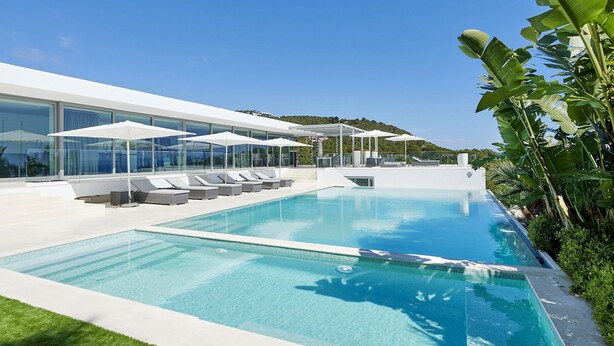 Un espace piscine moderne, doté de chaises longues et de parasols, entouré de murs de verre et de plantes tropicales.