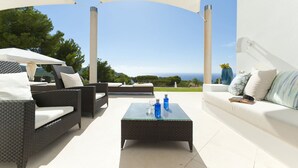 Un área de estar al aire libre con mobiliario de mimbre, una mesa con superficie de vidrio y vista al mar.