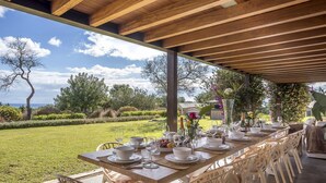Un espacio de comedor al aire libre con una larga mesa de madera dispuesta para una comida, rodeada de sillas blancas, con vistas a un jardín y árboles bien cuidados.