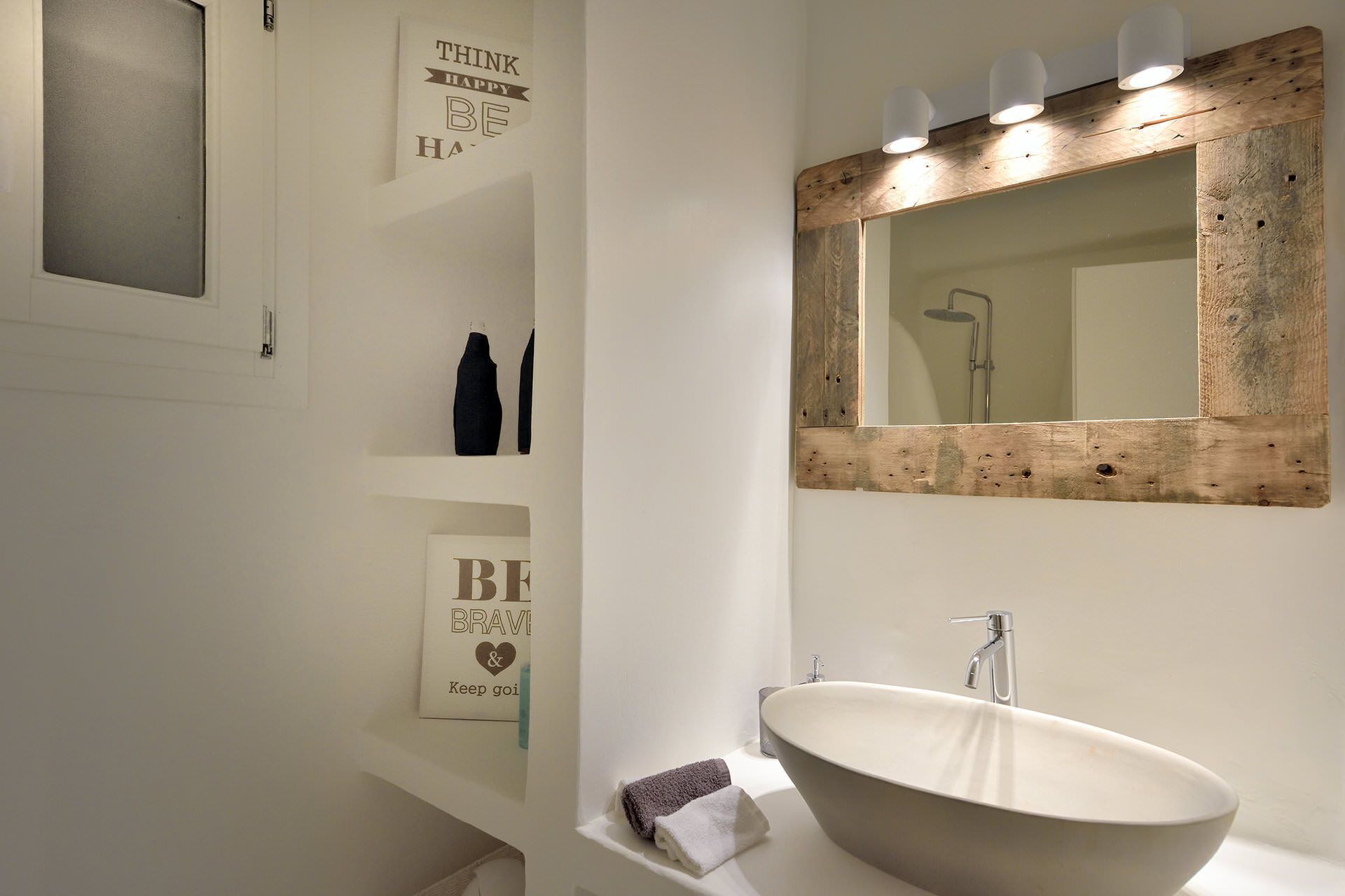 A modern bathroom with a large oval sink, a wooden-framed mirror, and a shelf with toiletries.