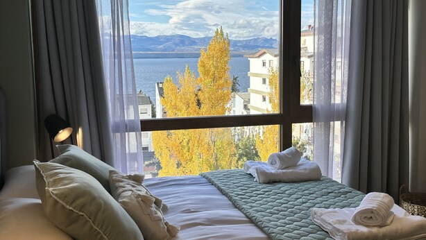 A hotel room with a bed, pillows, a quilted blanket, and a view of a lake and buildings.