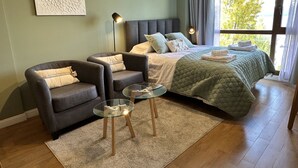 A modern bedroom with a bed, two armchairs, a glass coffee table, and a large window with curtains.