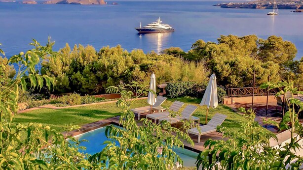 Zona de piscina con sillones de descanso y sombrillas, rodeada de vegetación y vistas a un mar tranquilo con yates.