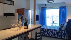 Een compacte kamer met kitchenette, een bed met beddengoed met een patroon, een eettafel met wijnglazen en een fles, en uitzicht op de omgeving door een groot raam.